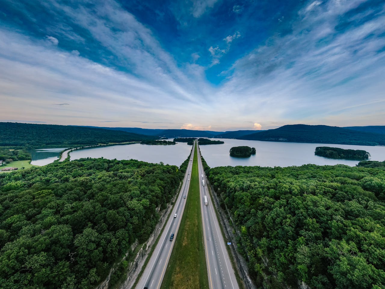 A breathtaking aerial shot of a highway running through lush forests beside a serene lake at sunset.
